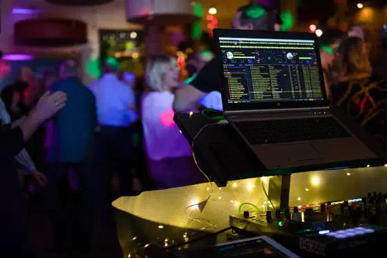 DJ-Pult mit Laptop und Controller, dahinter tanzende Gäste